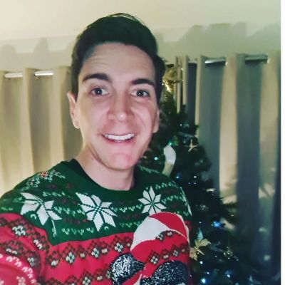 Oliver Phelps is wearing a sweater taking a selfie in front of a Christmas tree.